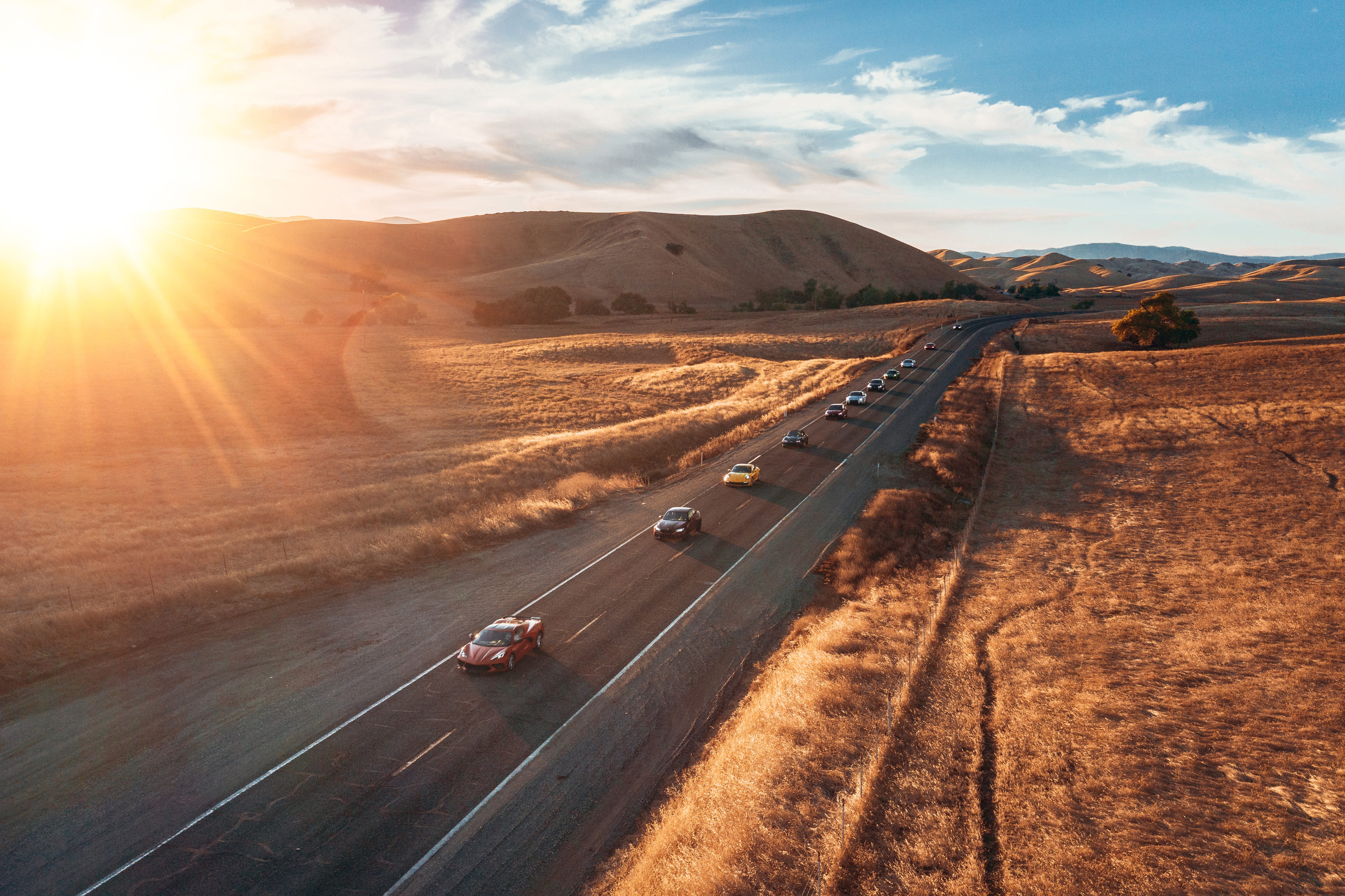 A line of luxury cars driving down a long road as the sun sets behind the mountains