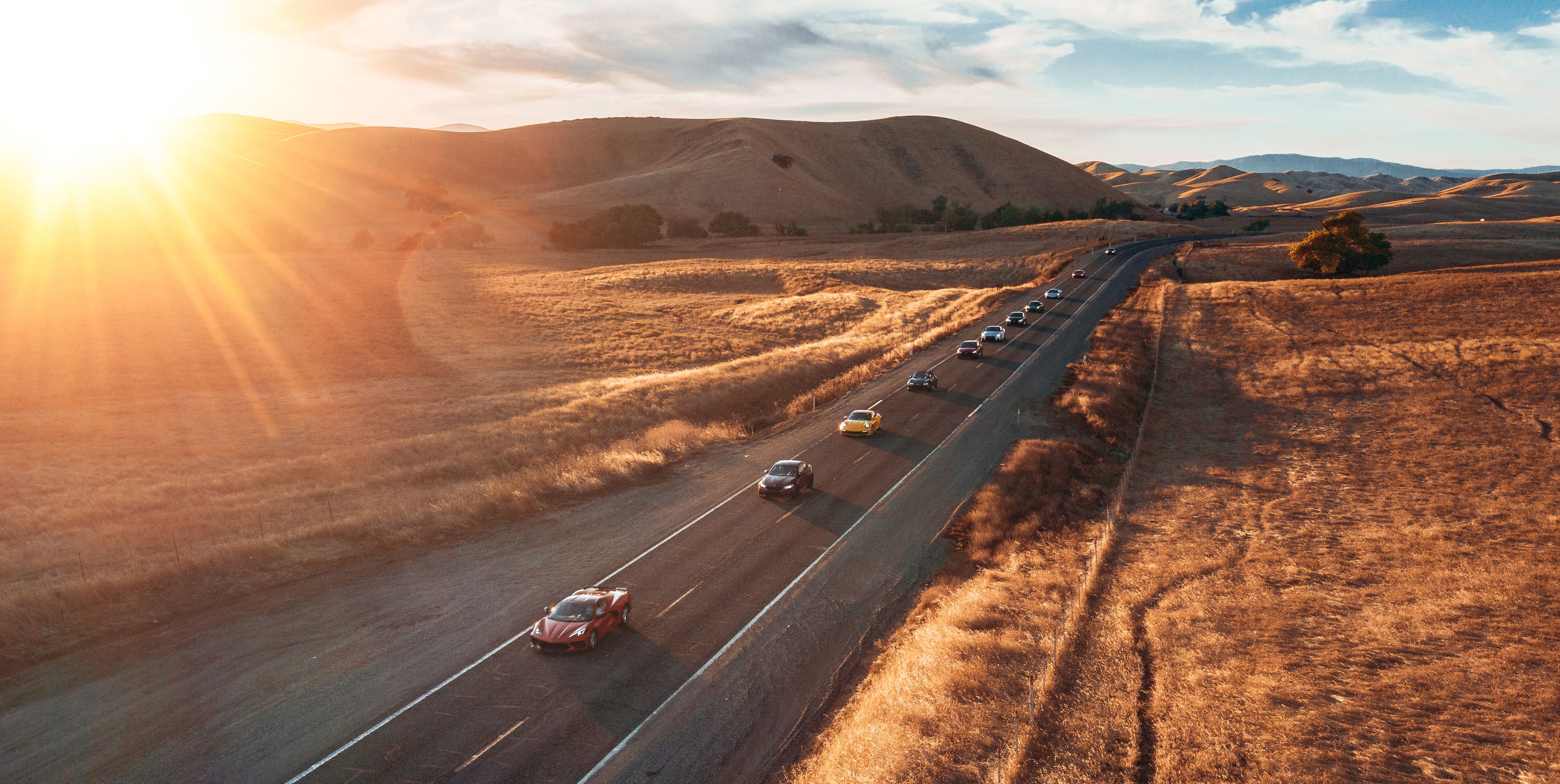 A line of luxury cars driving down a long road as the sun sets behind the mountains