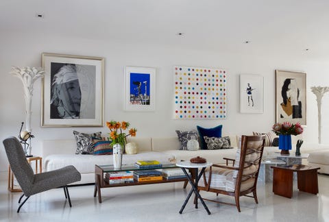 Caroline Rafferty - ELLE DECOR living room with glossy white interior, long white cushioned sofa along the wall and several chairs and low tables in front and five pieces of art hanging on the wall above the sofa, one of which is a large modernistic sketch of albert einstein on the far left