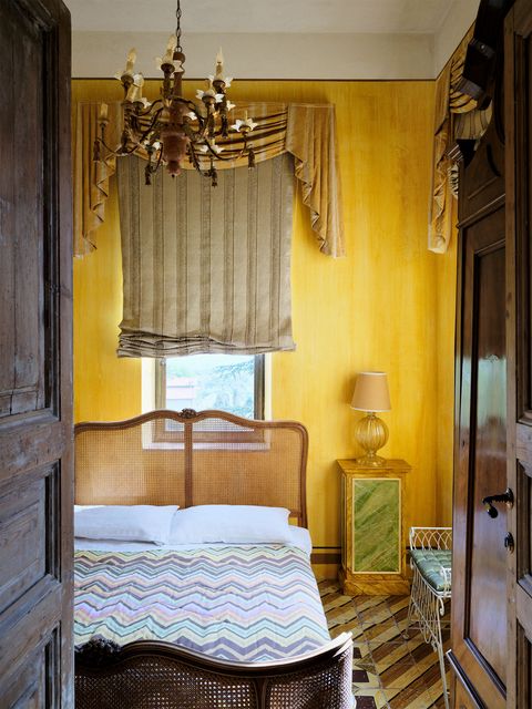 an ochre guest bedroom has a bronze chandelier, silk curtains and shade, a bed with caned head and footboards and multicolored zigzag patterned spread, a faux marble nightstand with a glass based lamp