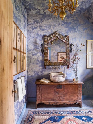 rustic powder room with patterned rug and a distressed wooden console holding a bowl sink and large mosaic mirror above it and tree patterene wallpper on the wall