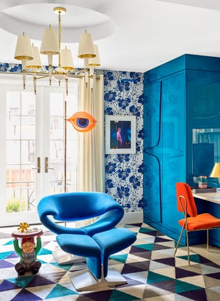 sitting room with a modern blue chair at center and a large shiny blue cabinet in the right corner a triangular patterned carpet in blues and taupes and the back wall has patterned flower wallpaper in blue