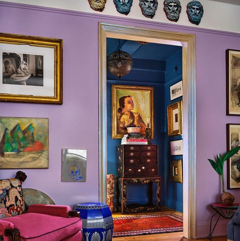 seating area with lilac walls looking out into another small room with darker blue walls and a highboy against the wall the carpet in the foreground is patterned purple and blue and the deep chair is red velvet with yellow fringed passementerie along the bottom