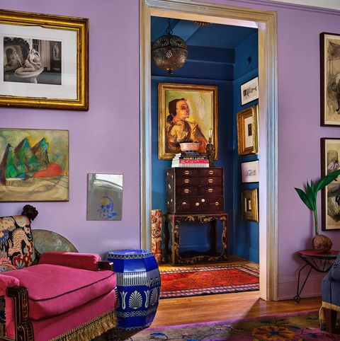 seating area with lilac walls looking out into another small room with darker blue walls and a highboy against the wall the carpet in the foreground is patterned purple and blue and the deep chair is red velvet with yellow fringed passementerie along the bottom