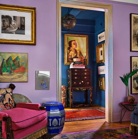 A List 2022 - ELLE DECOR seating area with lilac walls looking out into another small room with darker blue walls and a highboy against the wall the carpet in the foreground is patterned purple and blue and the deep chair is red velvet with yellow fringed passementerie along the bottom