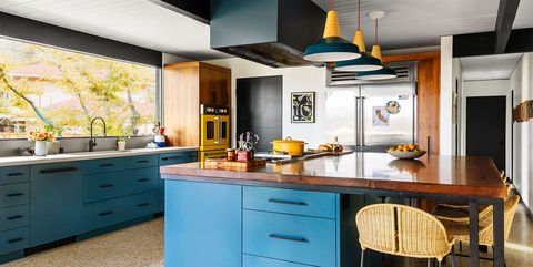 the kitchen has an island with cabinetry below a walnut slab countertop with six stools and pendants hanging above, a large window is above a sink and blue cabinets, a yellow oven is at the far end