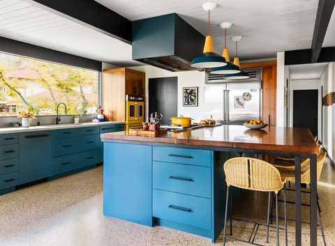 the kitchen has an island with cabinetry below a walnut slab countertop with six stools and pendants hanging above, a large window is above a sink and blue cabinets, a yellow oven is at the far end