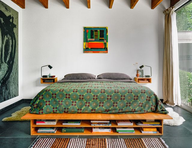 primary bedroom has artwork on the left and back walls, bed has suspended nightstands on both sides and a green print coverlet and dark gray linens, low wooden bookshelf at foot of bed and animal rugs on either side