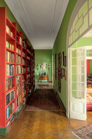 entry hall is lined with red painted bookshelves facing green walls hung with artworks, a turkish floor runner leads to an 18th century french commode at the far wall