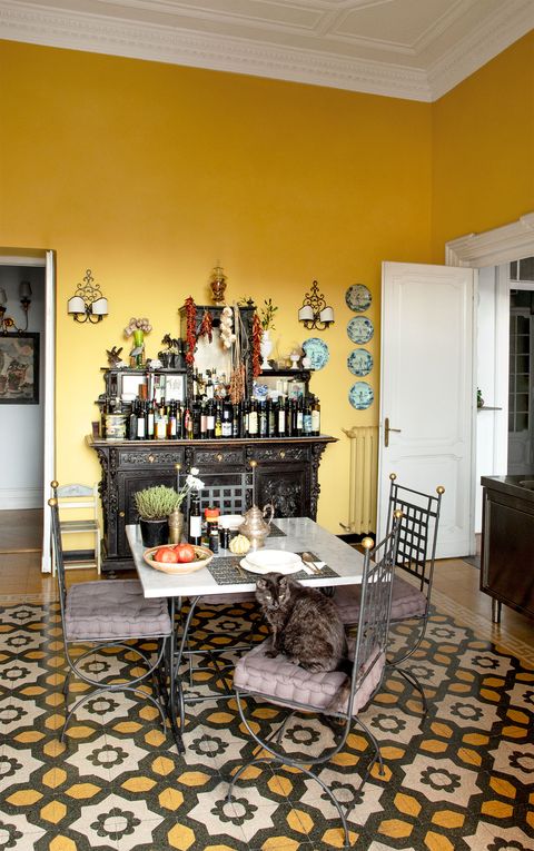 in the kitchen is a small dining table and metal chairs with cushions, cementine floor tiles, a black cabinet holding bottles and objects on top, and cat seated on one of the chairs
