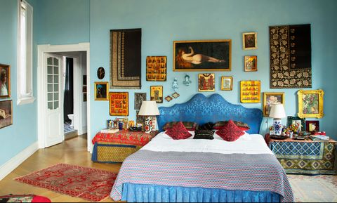 primary bedroom has pale blue walls with a king size bed with deep blue fabric headboard, side tables covered in embroidered fabric, small turkish rugs, and multiple artworks on the walls