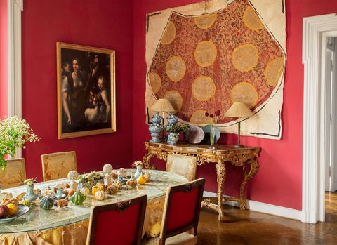 the dining room has red painted walls and a table with an embroidered silk tablecloth and 18th century chairs, a wall hanging is above an italian baroque console, and a painting hangs on the opposite wall