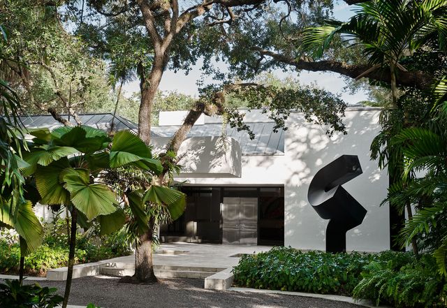 exterior of the modern home with a circular drive, a black steel sculpture, an oak tree in front of steps leading to a black set in entrance, with trees and shrubs surrounding the property