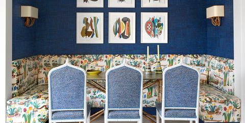 dining table set into an alcove where fabric cushioned chairs face a three sided banquette covered in a spring print fabric, two sconces and colorful framed artwork hang on deep blue walls behind the banquette