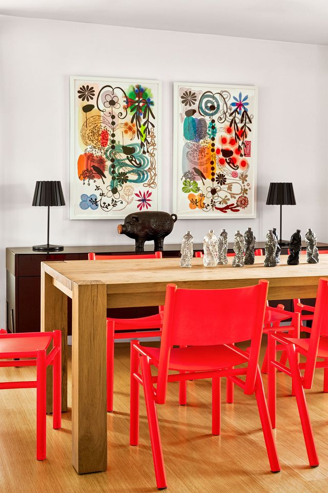 dining room has a wood table and on it are resin water bottles made to look like figures, red chairs, a console with two table lamps and a pig sculpture, two colorful abstract artworks hang above console