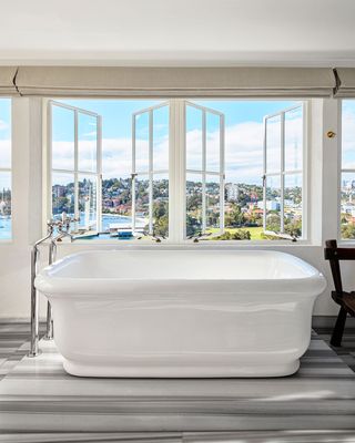 a deep bathtub, with fittings outside it, sitting on a raised marble platform in front of open windows that look out onto a harbor
