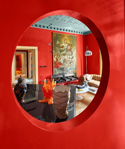 a view through a porthole from the dining room to the living room vases atop a baby grand piano, a leather sofa, cocktail table, two wassily chairs, and a large oil painting on the wall behind them