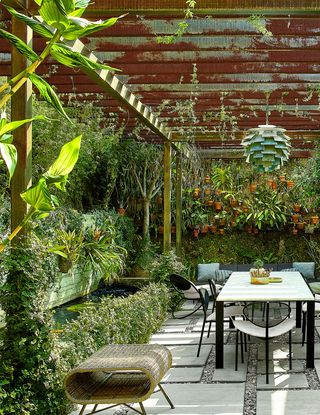 on an outside patio below a steel pergola is a cane bench, a dining table with chairs, and a pendant overhead, on the ground concrete tiles sit atop stone pavers, there is a koi pond and hanging pots with plants