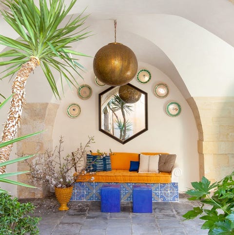 A List 2022 - ELLE DECOR covered outdoor area with orange daybed and bright blue stools and a mirror above and a semi circle of plates and giant orb hanging at center