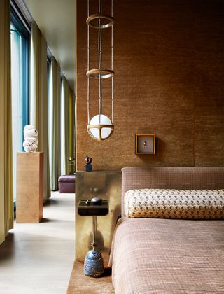 A copper bed with a fabric headboard and matching bedspread, the back wall is covered in an amber gold cloth, a marble and leather bedside table, and a copper, glass, and suede pendant light.