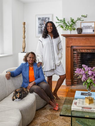 alyse archer coité beside her mother, sitting on the sofa with a calico cat, brick fireplace with wood surround and artwork on the mantel, a tall, thin wood carving on a pedestal in the corner