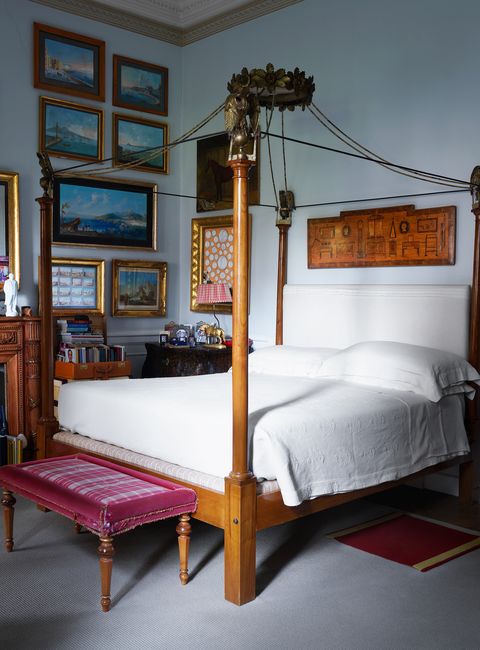 bedroom with pale blue walls, a wood canopy bed with eagles atop the columns and a white coverlet, antique bedside table with a lamp, bench with plaid fabric seat, at foot of bed, multiple framed artworks