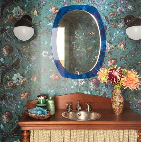 close up of a small wooden vanity with a yellow curtain and a small pitted stainless sink and green floral wallcovering behind it