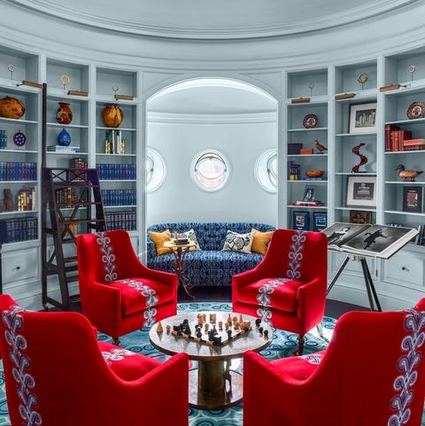 round room with four bright red velvet chairs in a circle around a small table shaped like a spool with chess board on it and a blue tufted banquette tucked into the far wall