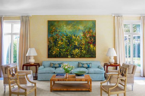 open airy living room with floor to ceiling french doors on either side of a light blue sofa with dark wood end tables on either end a low striated wood coffee table is in front as well as four chairs facing in a large floral painting hangs over the sofa