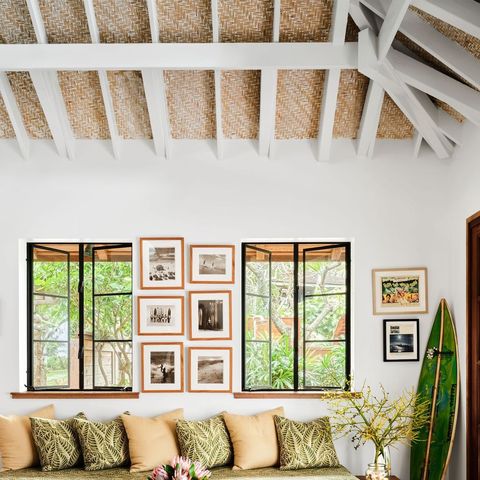 thin mattress upholstered in green palm fronds on a platform with many pillows and a low two tier table and two windows behind and multiple framed pieces on the wall