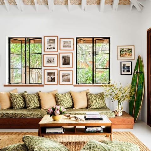 thin mattress upholstered in green palm fronds on a platform with many pillows and a low two tier table and two windows behind and multiple framed pieces on the wall