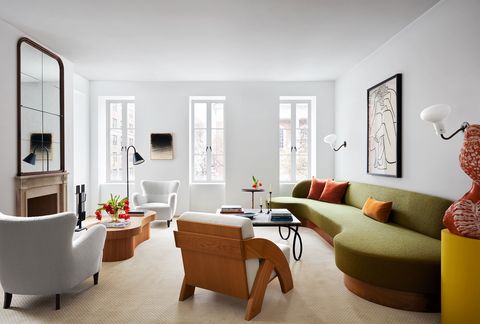 una sala de estar tiene tres ventanas, paredes blancas, una chimenea con una repisa tallada y un gran espejo arriba, dos sillones, un sillón, dos mesas de cóctel, un largo sofá curvo verde, apliques de pared y obras de arte