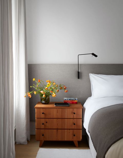a bed has a headboard of oatmeal that extends along the wall with a reading lamp attached, a side table with three drawers and books and a vase of flowers on top, curtains hanging on the window