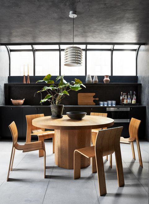 in a penthouse terrace room is a round wooden plinth table with five modern chairs and a light pendant, dark cabinets and shelves hold bottles, glass and candles, with arched windows above