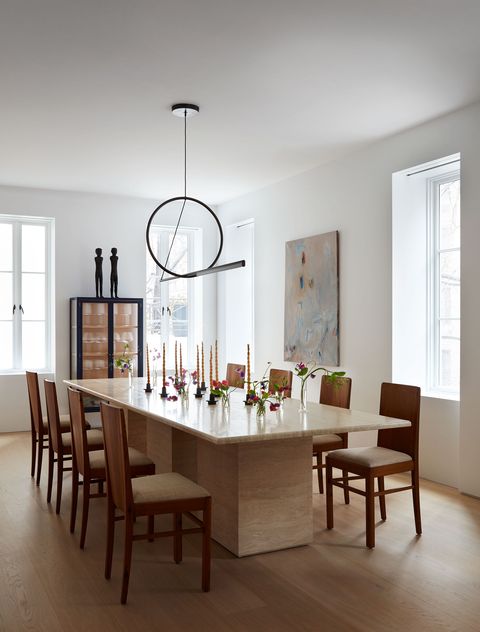 in a dining room, several candlesticks sit on a rectangular travertine table with eight chairs and a minimalist chandelier above it, a cabinet with glass doors and two sculptural figures upstairs and a work of art