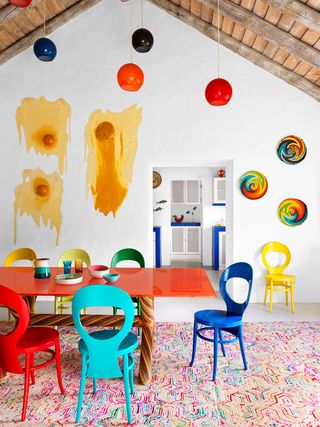 table à manger surmontée de verre orange avec des pieds en rotin tissé et des chaises peintes de couleurs vives, un tapis à motifs multicolores, cinq suspensions, des paniers lumineux à motif tourbillonnant et de l'art en résine accroché au mur