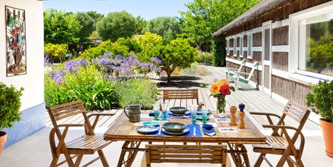 a covered terrace outside a wing of the house has a square bamboo dining table with four chairs, a deck with lounge chairs and small trees, tall grasses, and bushes with lavender colored blossoms