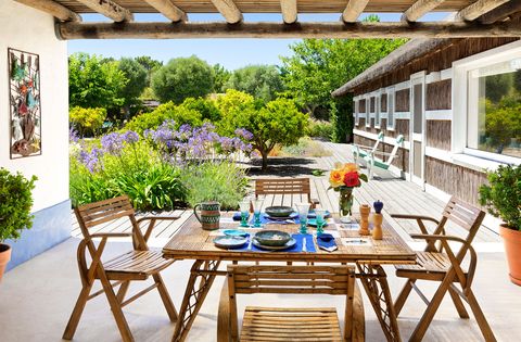une terrasse couverte à l'extérieur d'une aile de la maison a une table à manger carrée en bambou avec quatre chaises, une terrasse avec des chaises longues et de petits arbres, de hautes herbes et des buissons avec des fleurs de couleur lavande