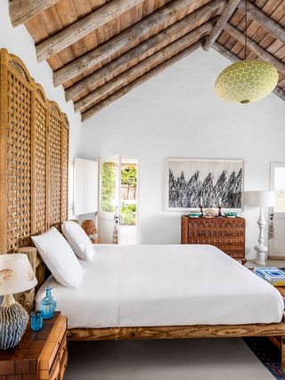 main bedroom with rustic wooden bed frame and large rattan headboard, white linens, wooden side table with drawers and lamp with a shell motif, a mahogany cabinet, a pendant light, and a painted white floor lamp