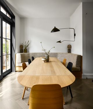 dining area has a wood frame curved banquette with beige patterned fabric seating tucked into a nook, an extendable wooden table with alternating natural oak and black ash chairs, a sconce with two lights on wall