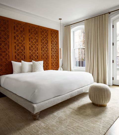 primary bedroom has a wood and linen fabric bed frame with white linens, moorish carved wood doors line the wall behind bed forming a headboard, the drapes, a beige pouf, and the rug are beige colored