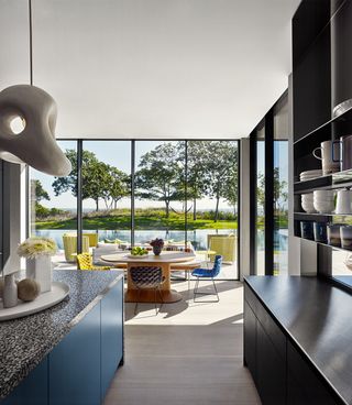 a kitchen has a wall of glass overlooking a swimming pool, steel blue floor cabinets with a terrazzo countertop opposite dark wood cabinets with a steel countertop with shelving above, a round dining table with terrazzo chairs woven fabric in different colors