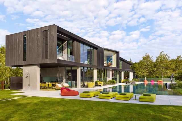 the exterior of the two story house with charred wood cladding on the upper floor and glass walls and concrete on the lower looks out onto a large swimming pool and deck with bright green, pink, and orange lounge chairs