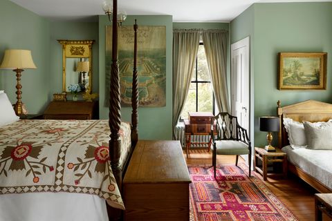 a bedroom has muted green walls, a four poster bed with a rose motif quilt, a slant top desk in a nook, a trunk, a wall tapestry, a luggage stand with antique cases, a curtained window, and a patterned runner