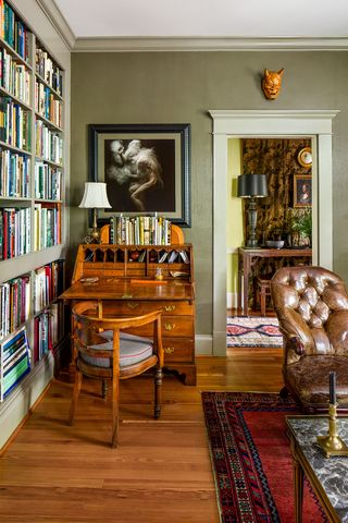 una biblioteca tiene un escritorio chippendale con cajones y compartimentos pequeños, libros y una lámpara en la parte superior, una foto grande enmarcada arriba, una silla con respaldo curvo en el frente, estanterías, una silla de cuero y una alfombra roja estampada