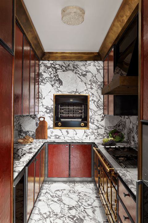 a galley kitchen has cabinets below and above counters that have cracked red lacquer, the floor, counters, and walls are white and black marble, a rotisserie is on the back wall, a stove and oven are at right with a sink at left
