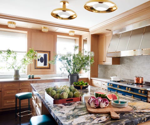 cocina con una gran isla cubierta de mármol cubierta de comida en primer plano y estufa azul la cornue a la derecha y elegantes gabinetes de madera modernos en el fondo