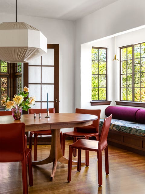 oval double pedestal wood table with snazzy and sleek red leather armless chairs pulled up to it and in the right background is a built in upholstered bench with windows behind it