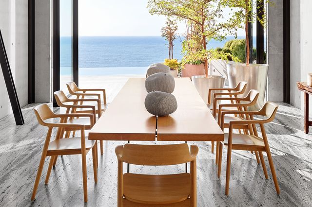clean spare dining room with a view of a long light wood dining table and chairs with rock like sculptures on it and gray streaked floor with black and white and globe suspected lighting above the table all opening out from floor to ceiling glass doors to a glorious view of the water