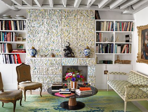 living room area with large tiled wall with inset for a planter and built in bookshelves on either side and some chairs and a settee in a chain link pattern upholstery and a round low pedestal table at center heaped with books on a green rung